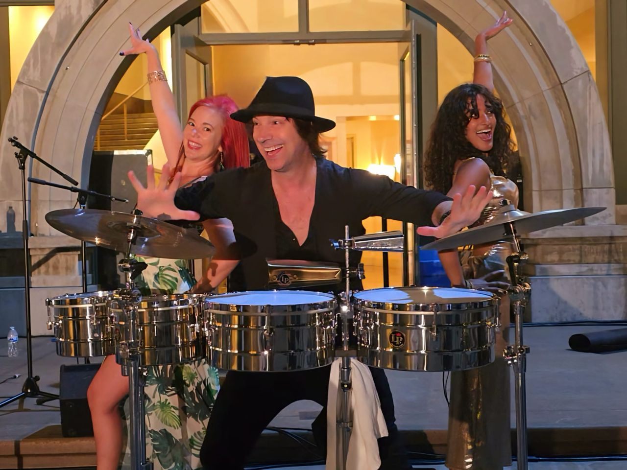 Michael John Saltus on timbales with two Positive Energy dancers performing behind him at an outdoor evening event